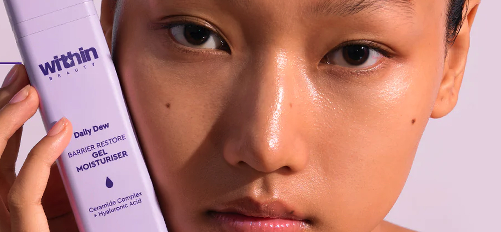 Close-up of a person holding a lavender bottle of Within Beauty Daily Dew Barrier Restore Gel Moisturiser next to their face.