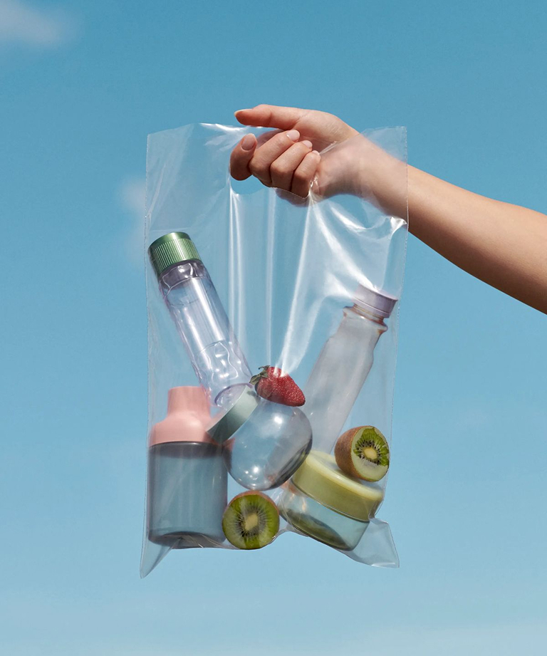 Hand holding a transparent bag filled with bottles & containers, along with kiwi slices and a strawberry, against a blue background.