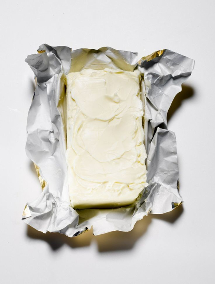 Block of butter partially unwrapped from silver foil packaging on a white background.