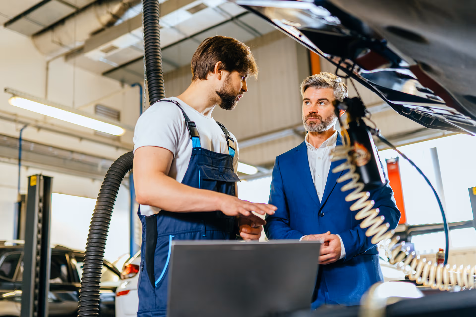Mechanic in overalls explaining car diagnostics to a man in a suit in an auto repair shop.