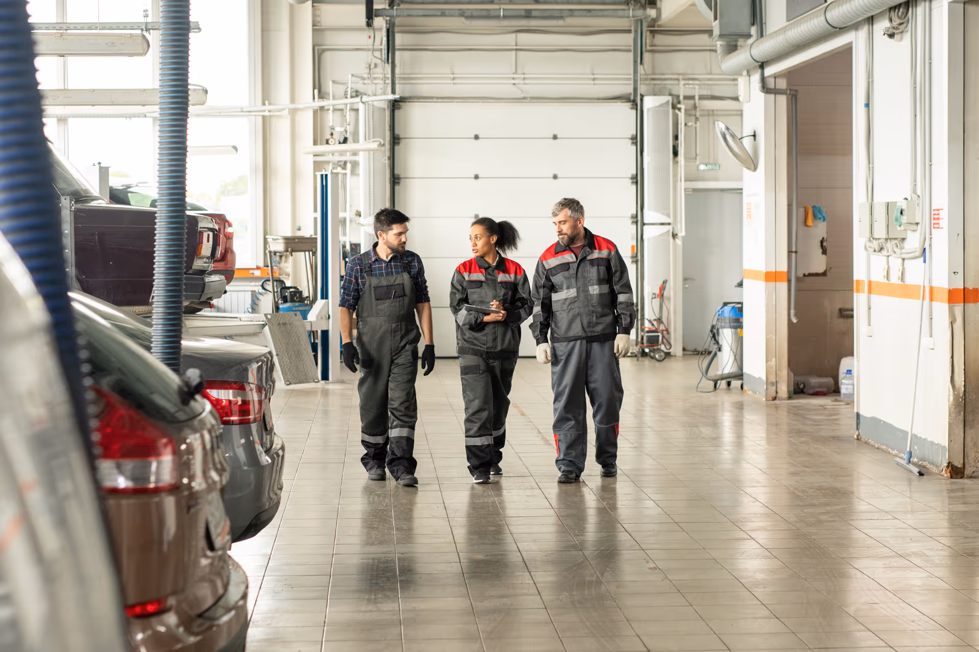 Three mechanics walking and talking inside a car repair garage with cars parked on one side.