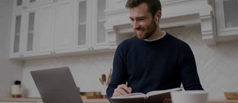 Un homme dans sa cuisine, devant son ordinateur, livre et stylo en main, écouteurs aux oreilles, représentant la diversité des lieux d’apprentissage et l’accessibilité des cours en ligne ouverts à tous.