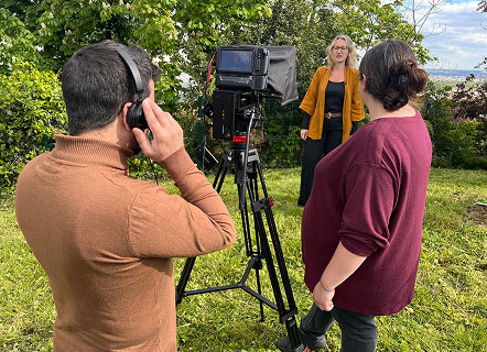 Deux professionnels, vus de dos, sont en pleine préparation d’une séquence vidéo pour un reportage. À l’arrière-plan, une femme participe à la dynamique de l’équipe, offrant une atmosphère de collaboration et de rigueur qui caractérise la réalisation de contenus audiovisuels de qualité.