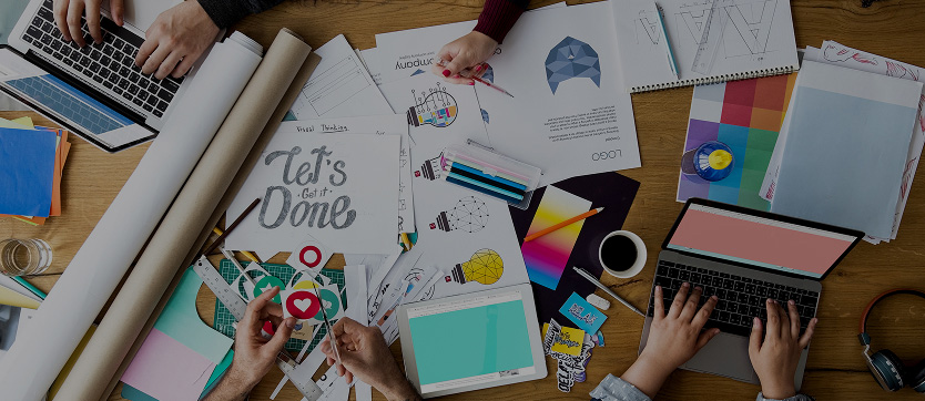 Vue aérienne d’une table de travail où une équipe de communication élabore un concept : rouleaux de papier, ordinateurs, tablette, pictogrammes, croquis de logo, nuancier et dossier de projet. L’image illustre la dynamique collective, la multiplicité des outils graphiques et numériques, et la phase d’idéation où chaque support contribue à la construction d’une identité visuelle cohérente et innovante.