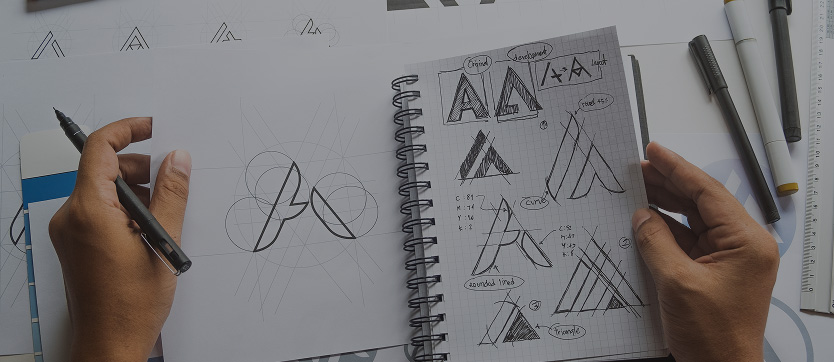 Vue du bureau d’un graphiste en pleine phase de création : une main gauche esquisse au crayon différentes idées de logo dans un carnet de croquis. Cette image symbolise la première étape du processus créatif, où l’inspiration, l’expérimentation et la multiplicité des pistes graphiques sont essentielles pour aboutir à un logo unique et pertinent.