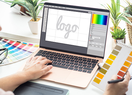Image illustrant la création graphique : un ordinateur sur une table montre un logiciel de design avec une ébauche de logo (le mot « logo »), tandis qu’un nuancier Pantone à proximité symbolise l’importance du choix des couleurs dans la conception d’identité visuelle.