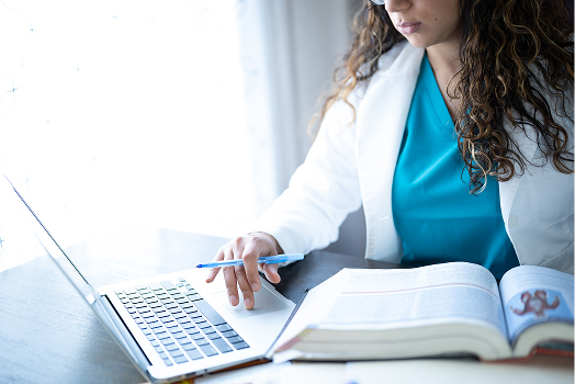 Femme en blouse médicale étudiant devant un ordinateur avec un manuel de médecine, suivant une formation en ligne
