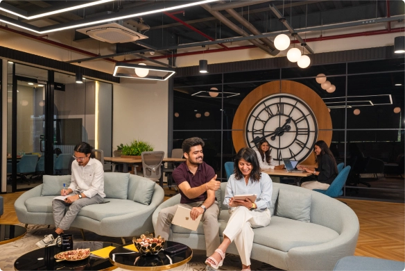People sit and work in a DevX Gift city modern office lounge with large sofas, a round coffee table, and a big wall clock. Some are using laptops or tablets, and the space has stylish lighting and decor.