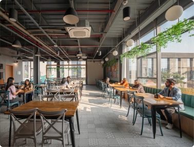 A modern, spacious café with large windows, several people sitting at tables, some conversing and others working alone; sunlight streams in, and green plants hang near the windows at DevX Gift city.