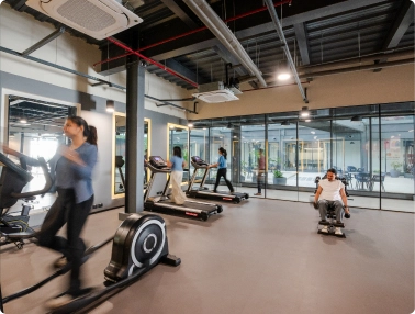 People exercising on cardio machines in a DevX Gift City modern gym with large windows and gray flooring.