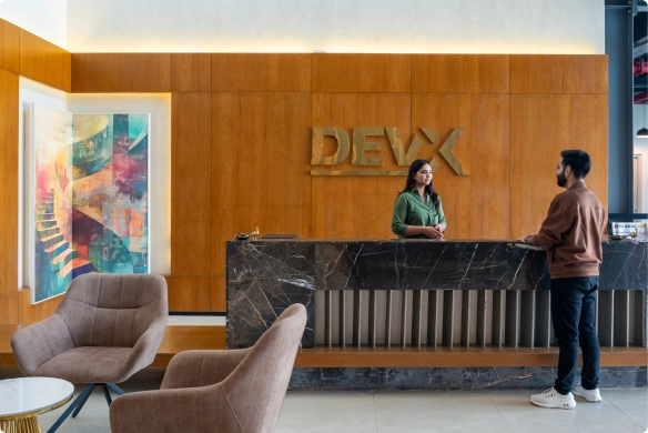 A woman stands behind a marble reception desk with a DEVX sign, speaking to a man at DevX Gift City; two modern chairs and a colorful abstract painting are visible.