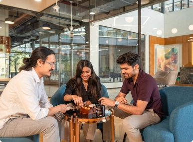 Three people seated around a small round table in an office lounge, playing a board game together at DevX Gift City.