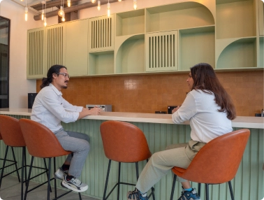 Two people sitting on orange chairs at a counter in a modern cafe with green and brown decor at DevX Gift City.