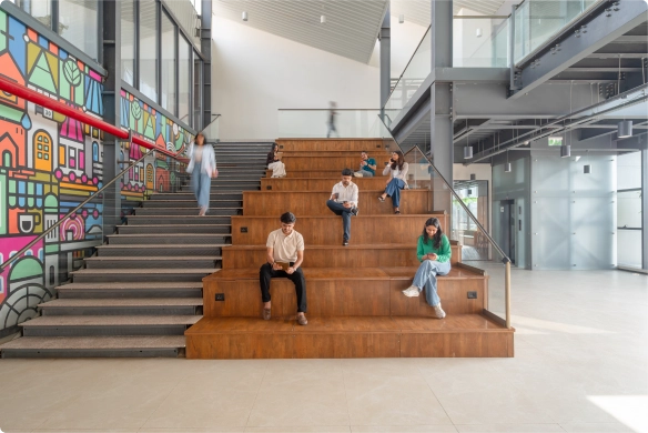 Five people sit separately on wide wooden steps in a DevX Gift city modern, spacious building with glass walls and a colorful mural on the adjacent staircase. The space is bright and open, with natural light streaming in.