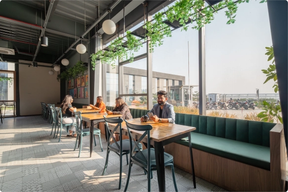 Modern cafe with large windows, green plants hanging from the ceiling, people sitting at wooden tables, and natural light filling the space at DevX Gift City.