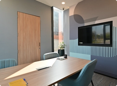 DevX Gift City modern meeting room with wooden table, blue chairs, wall-mounted TV, and sunlight from tall window.