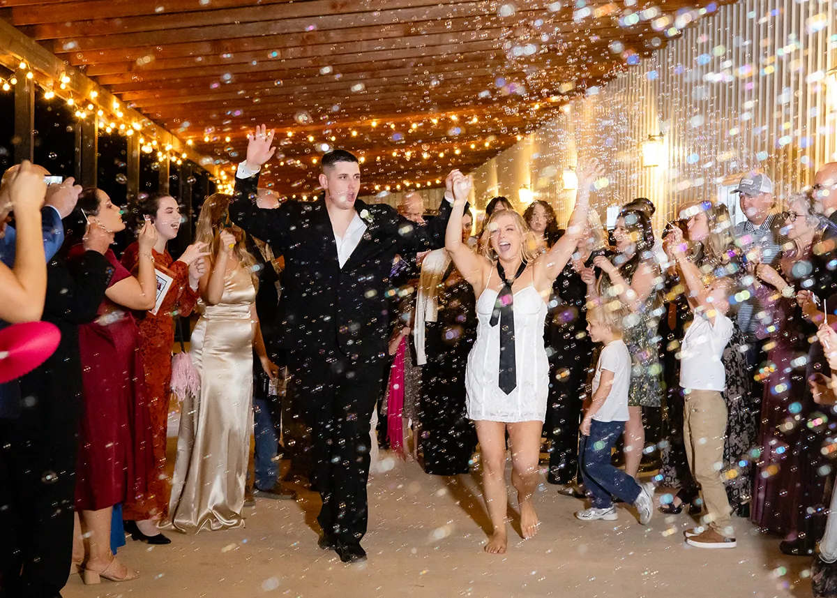 A newly married couple holding hands exiting their wedding reception while their guests line up blowing bubbles.