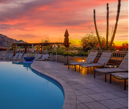 Empty poolside lounge chairs and umbrellas at sunset with colorful sky and desert plants nearby.