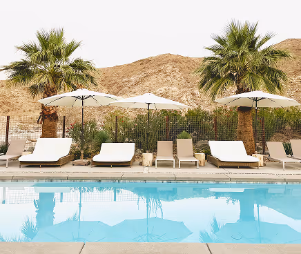 Poolside lounge chairs and umbrellas with palm trees and desert hills in the background.
