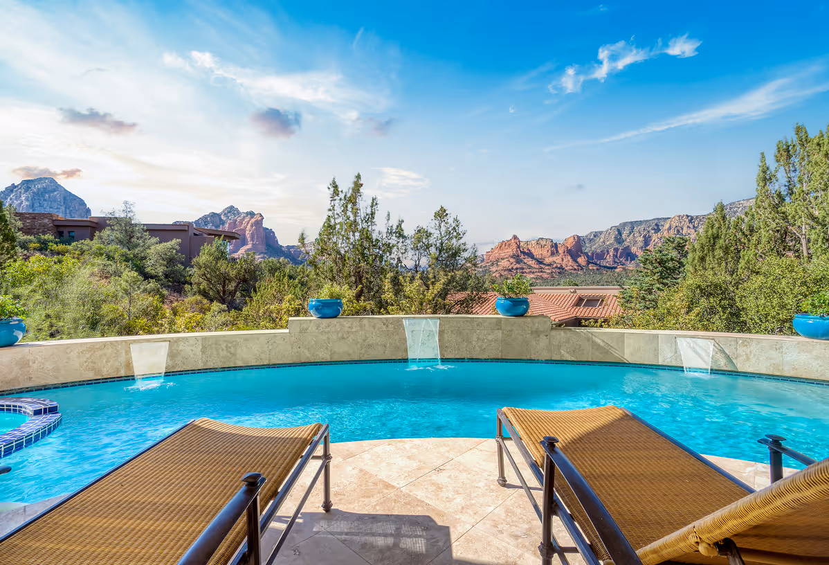 Swimming pool with water spouts and lounge chairs overlooking trees and red rock mountains under blue sky.