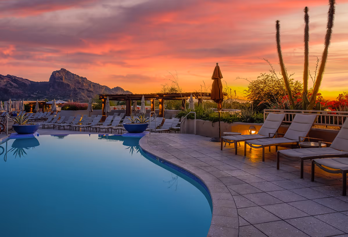 Outdoor pool with empty lounge chairs and umbrellas at sunset with mountains in the background.