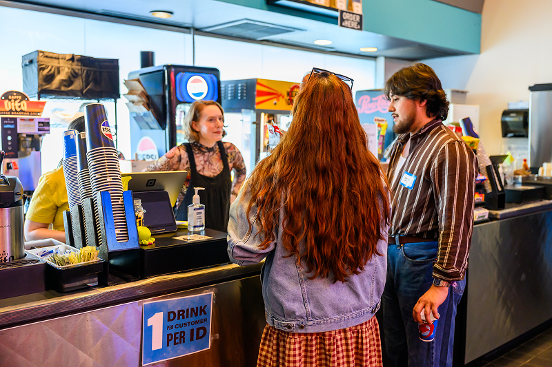 Two customers talking to a cashier at a drink counter with cups, soda machines, and hand sanitizer visible.