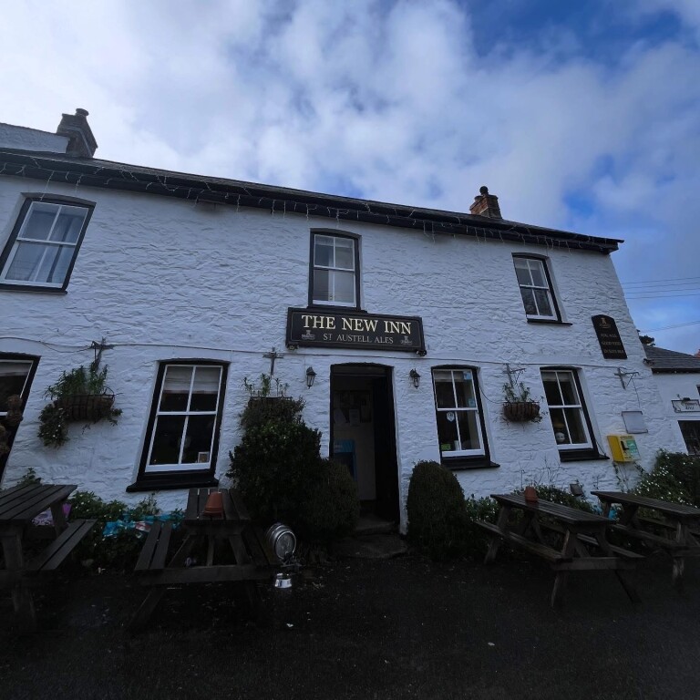 White stone building of The New Inn with black trim, hanging plants, picnic tables outside, and a cloudy blue sky.