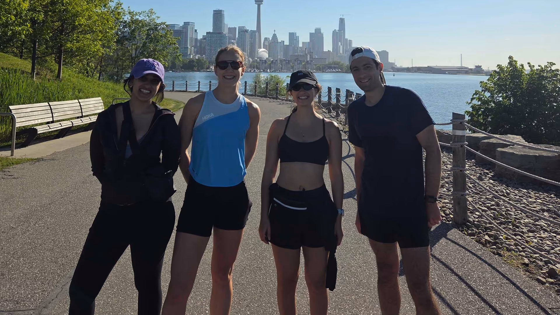 Four people in athletic wear standing on a waterfront path with a city skyline in the background.