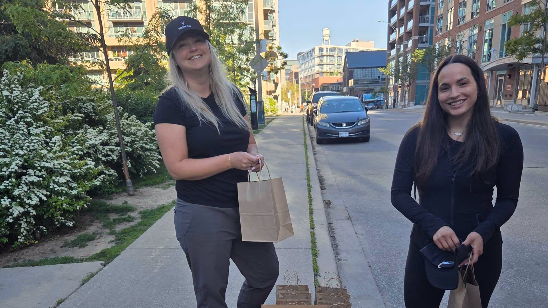 Two smiling women holding paper bags on a city sidewalk with buildings and cars behind them.