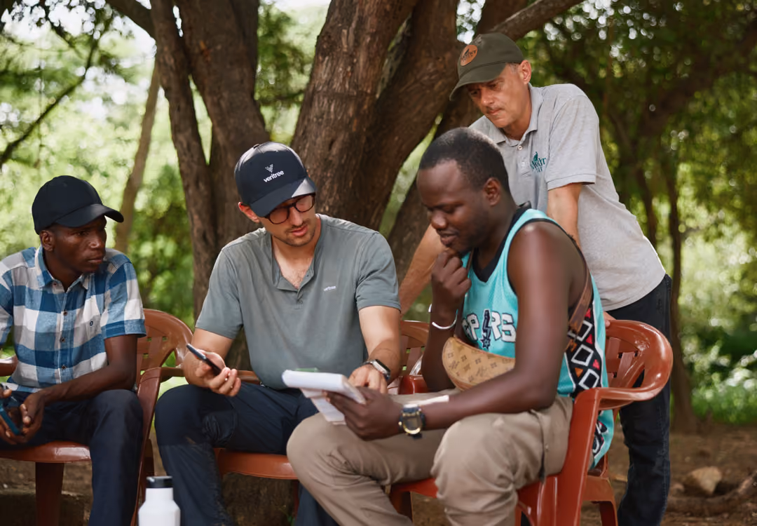 Four men sitting outdoors under trees, engaged in discussion with notepad and pen.