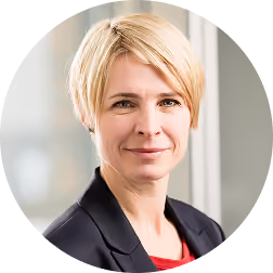 Professional headshot of a woman with short blonde hair wearing a black blazer and a red top, with a neutral expression and a blurred office background.