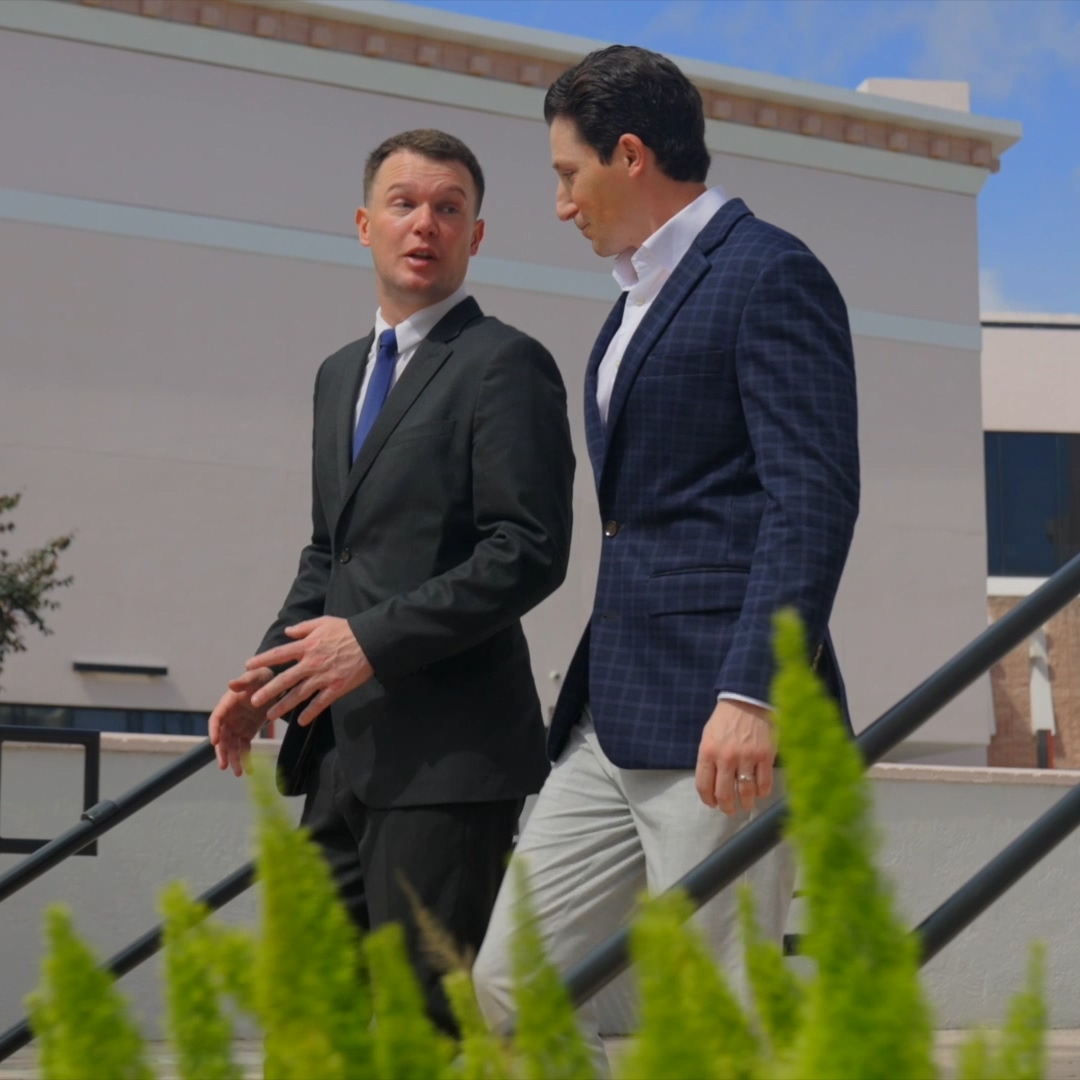 Two men in suits walking outdoors engaged in conversation near stairs and green plants.
