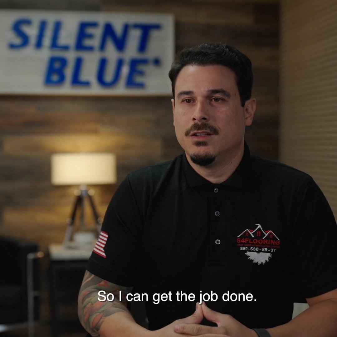 Man with tattooed arm in black shirt with a flooring logo speaks in an office with 'SILENT BLUE' sign.