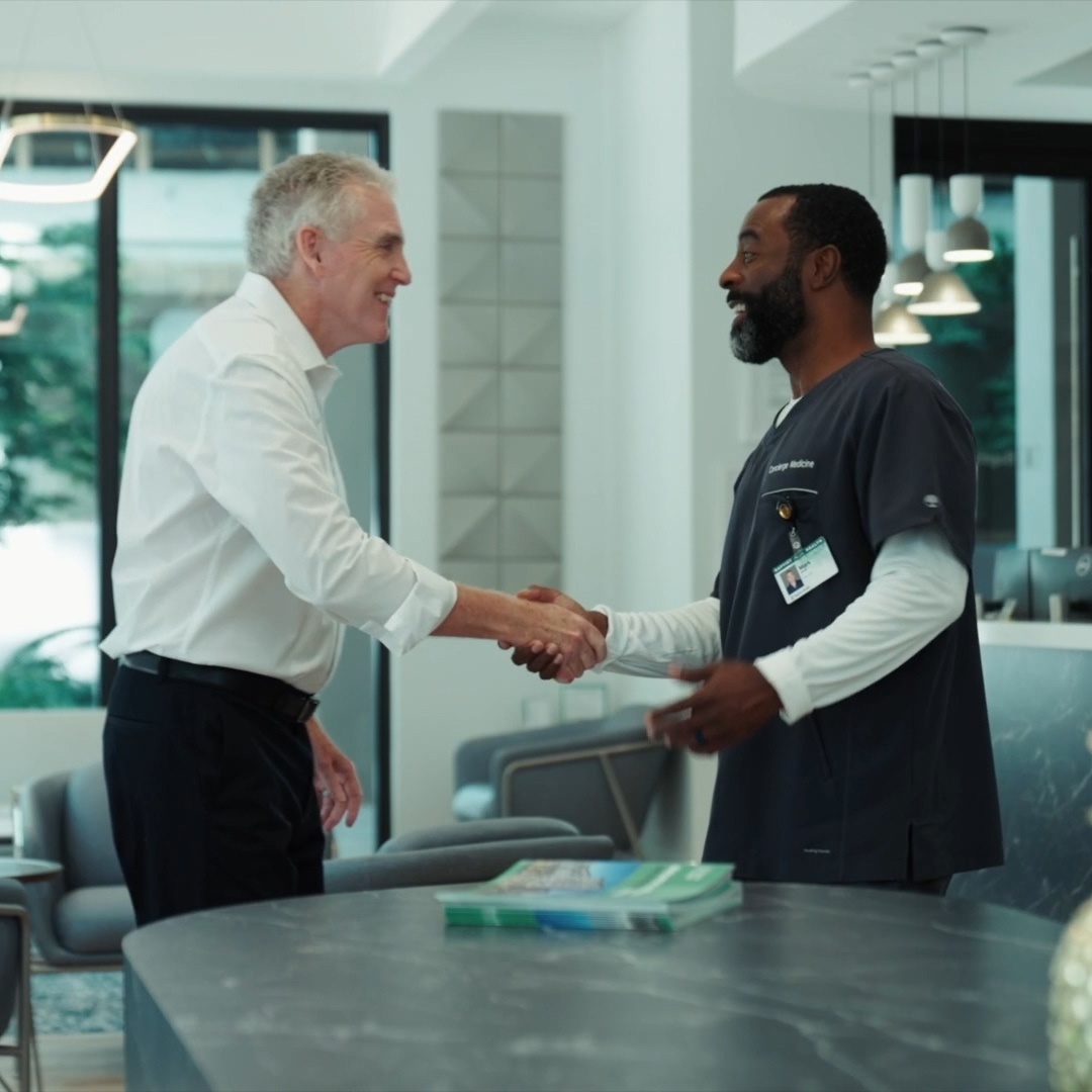 Smiling man in white shirt shaking hands with man in medical scrubs at reception desk.