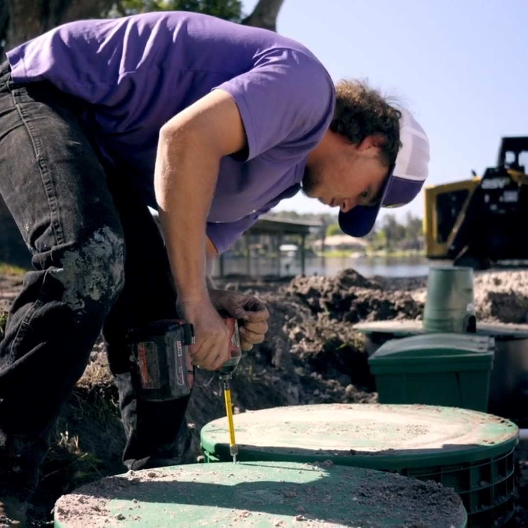 Worker in purple shirt using power drill on green utility covers outdoors near water.