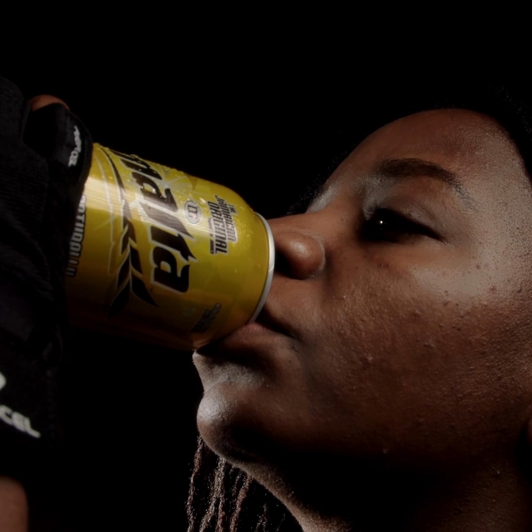 Person with braided hair drinking from a yellow can labeled 'TAYLOR' against a black background.