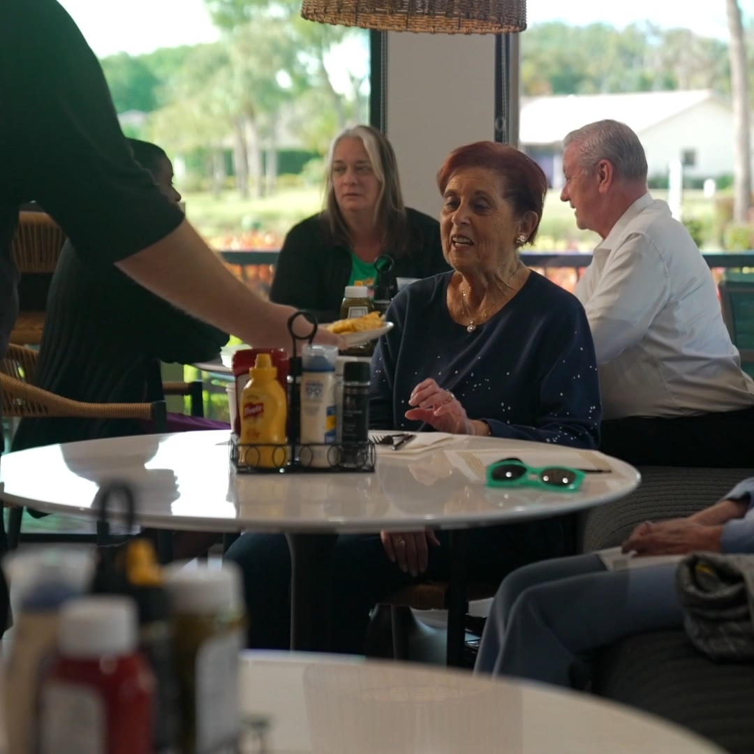 Elderly woman seated at a restaurant table receiving a plate of food from a server.
