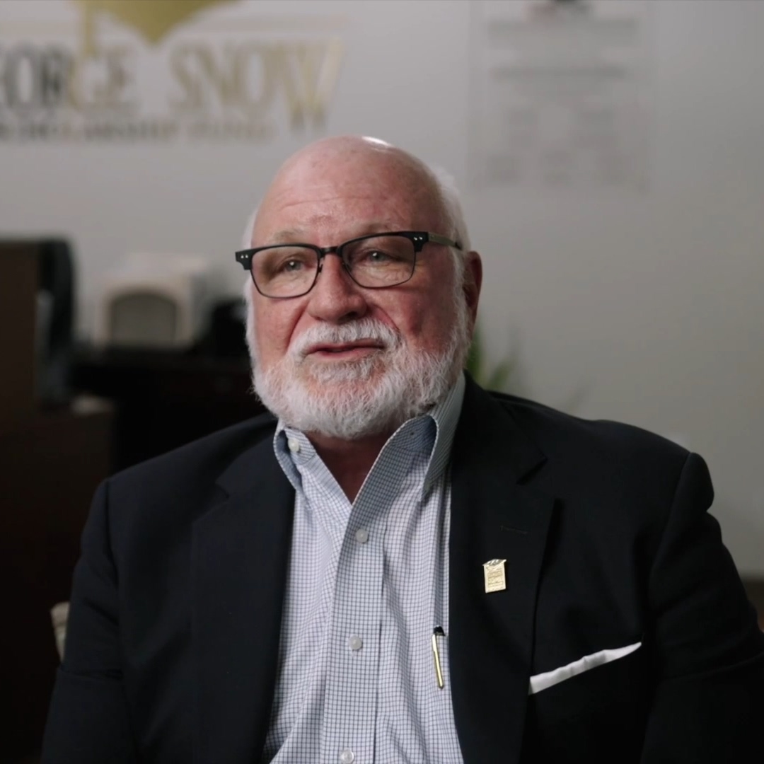 Elderly man with glasses, white beard, wearing a checked shirt, dark suit, and pin seated indoors.