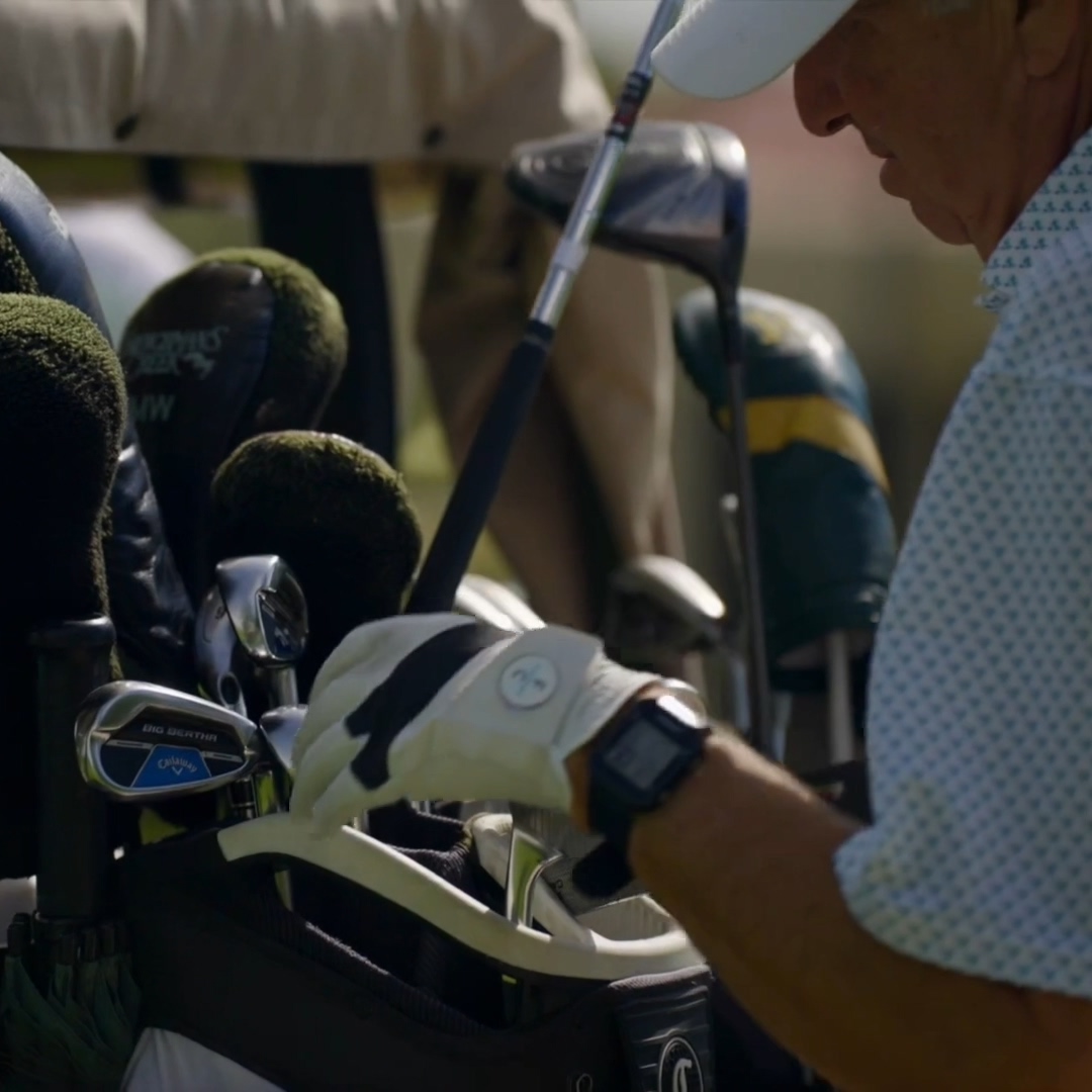 Man wearing golf glove reaching for a club in a golf bag filled with clubs and headcovers.