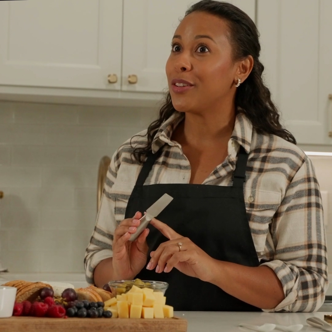Woman in plaid shirt and black apron holding a cheese knife with a charcuterie board in front of her.