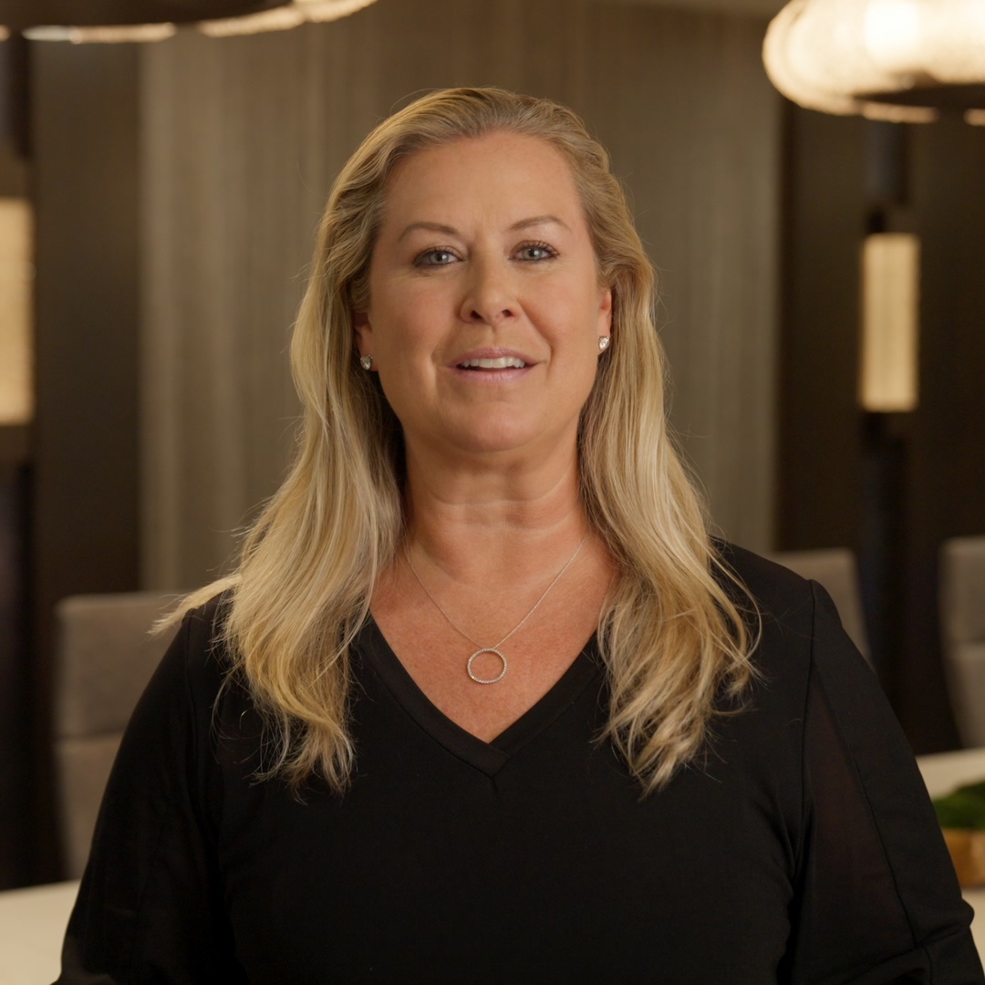 Smiling blonde woman with wavy hair wearing black top and silver necklace in a softly lit room.