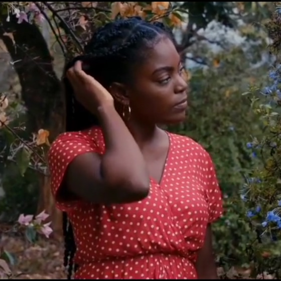 Woman in red polka dot dress adjusting braided hair in a garden with trees and flowers.