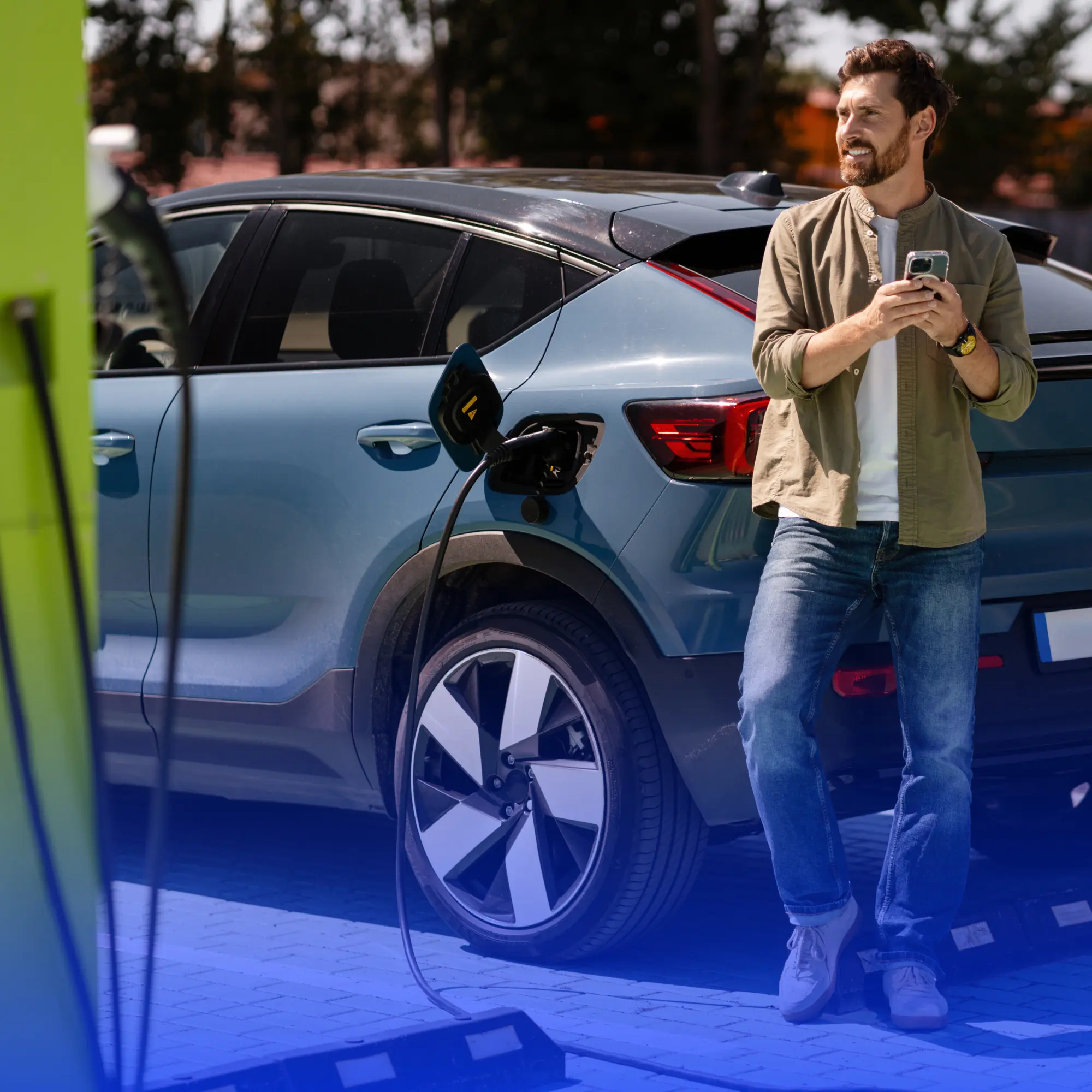 Homem sorrindo com celular na mão ao lado de carro elétrico sendo carregado em estação pública ao descobrir como comprar carro elétrico no Brasil