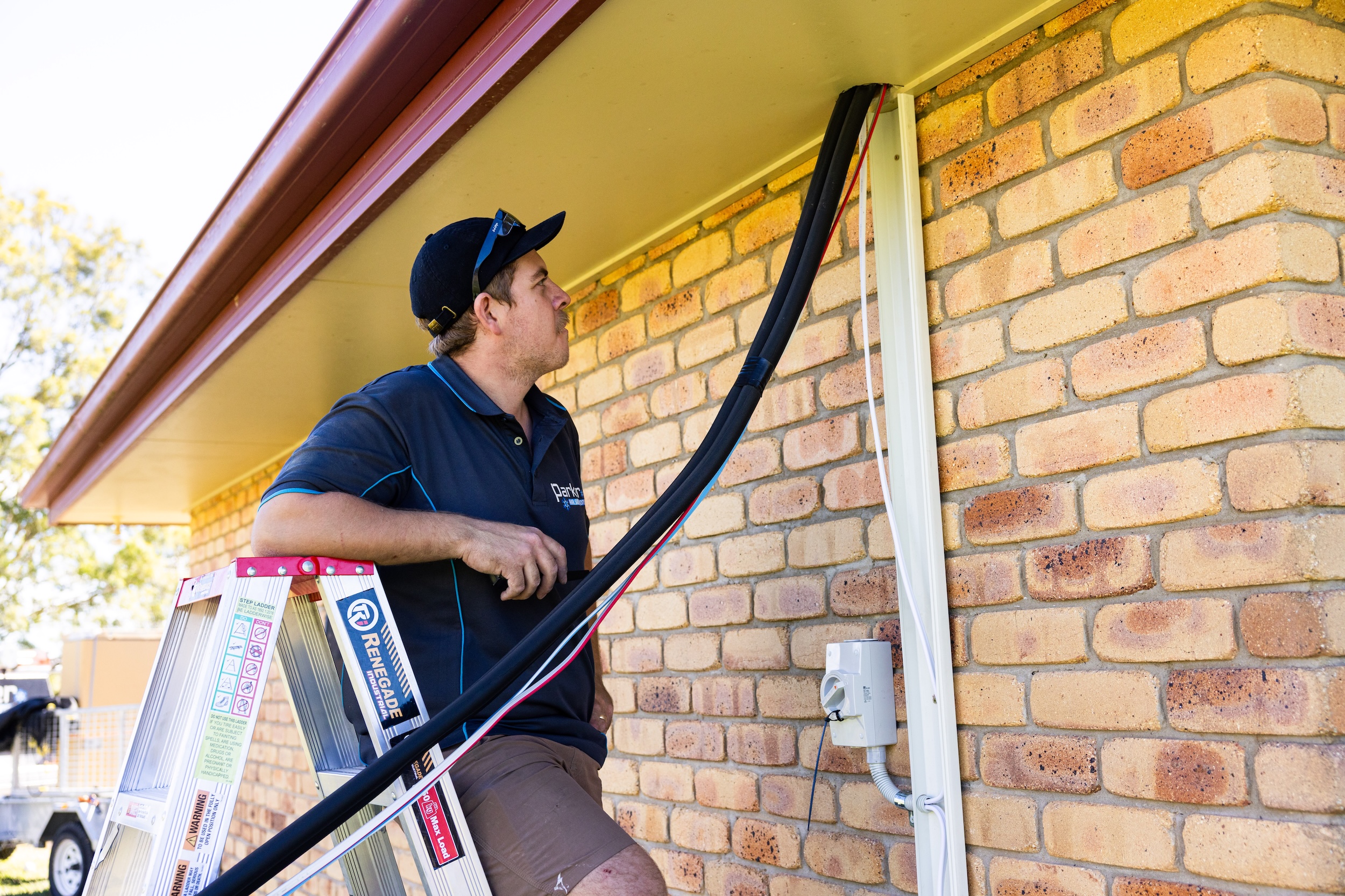 Professional installer on a ladder mounting black insulated pipes and white protective duct cover to a brick exterior wall.
