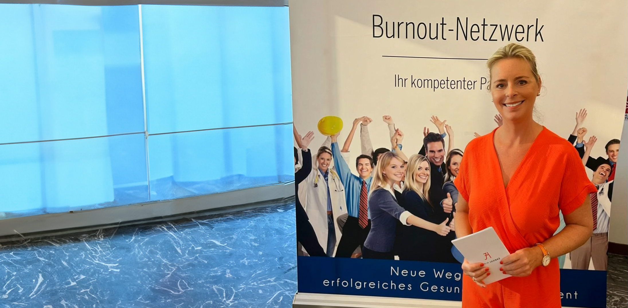 Lächelnde Frau in einem orangefarbenen Jumpsuit, die Notizzettel in der Hand hält und vor einem Plakat des Burnout-Netzwerks steht.