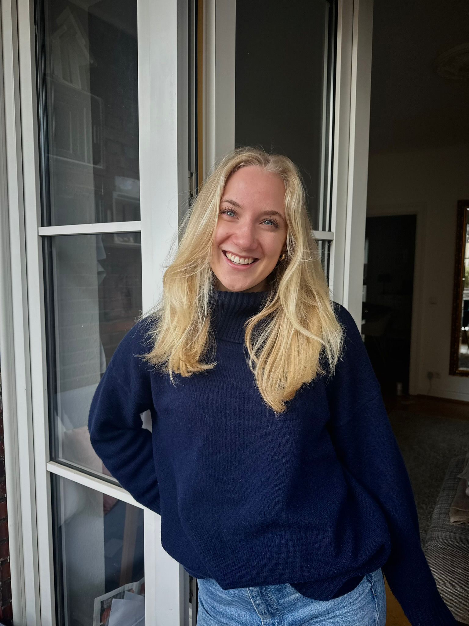 Frau mit blonden Haaren und blauem Sweatshirt steht in der Glastür einer Wohnung und lächelt.