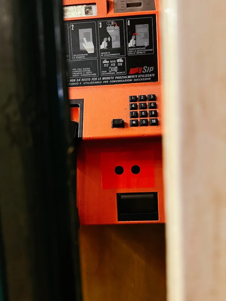 An orange Italian SIP payphone with the two-dot Odd Universe eyes on its coin box