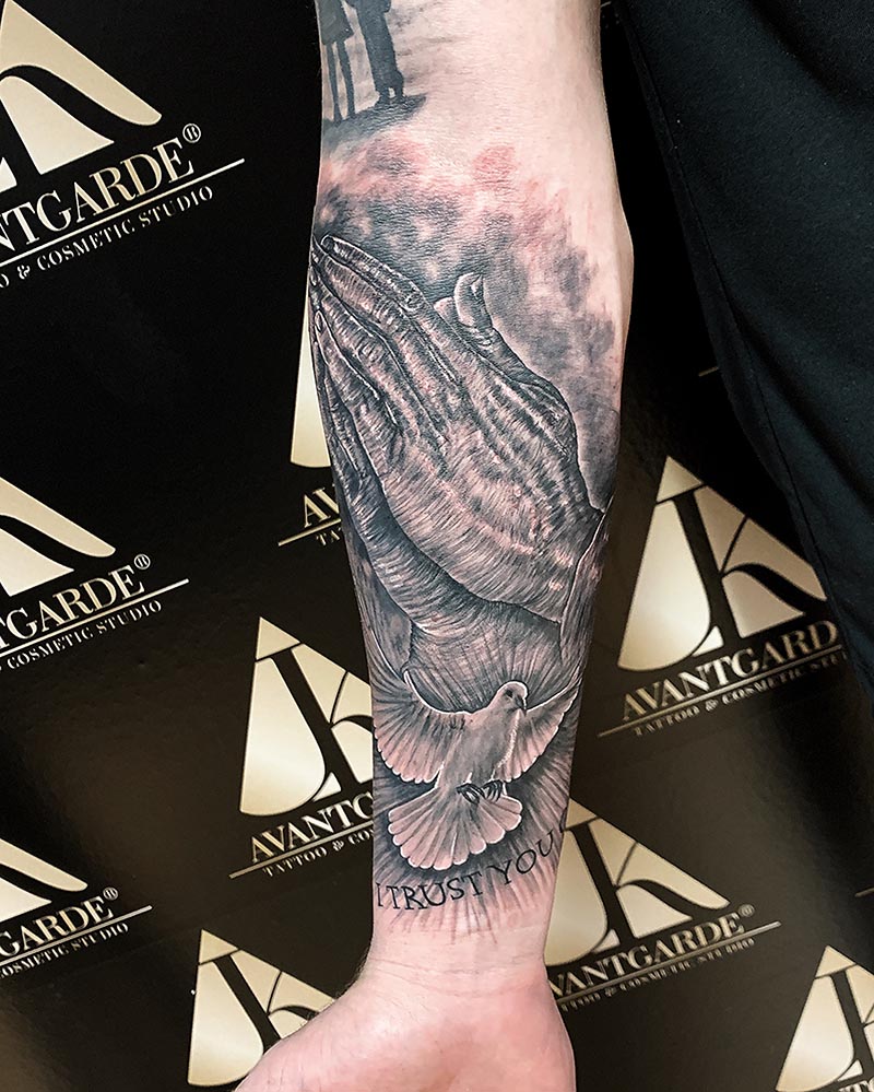 Schwarzgraues Tattoo mit Vogelkopf, Federn und Blüten am Unterarm.