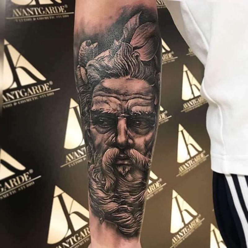 Schwarzgraues Tattoo mit einem bärtigen Gesicht und ornamentalen Formen am Arm.