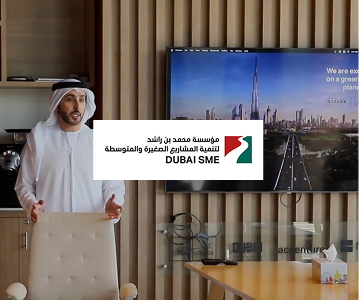 Man in traditional Emirati clothing standing beside a chair in a modern office, with a large screen displaying Dubai skyline and Burj Khalifa in the background.